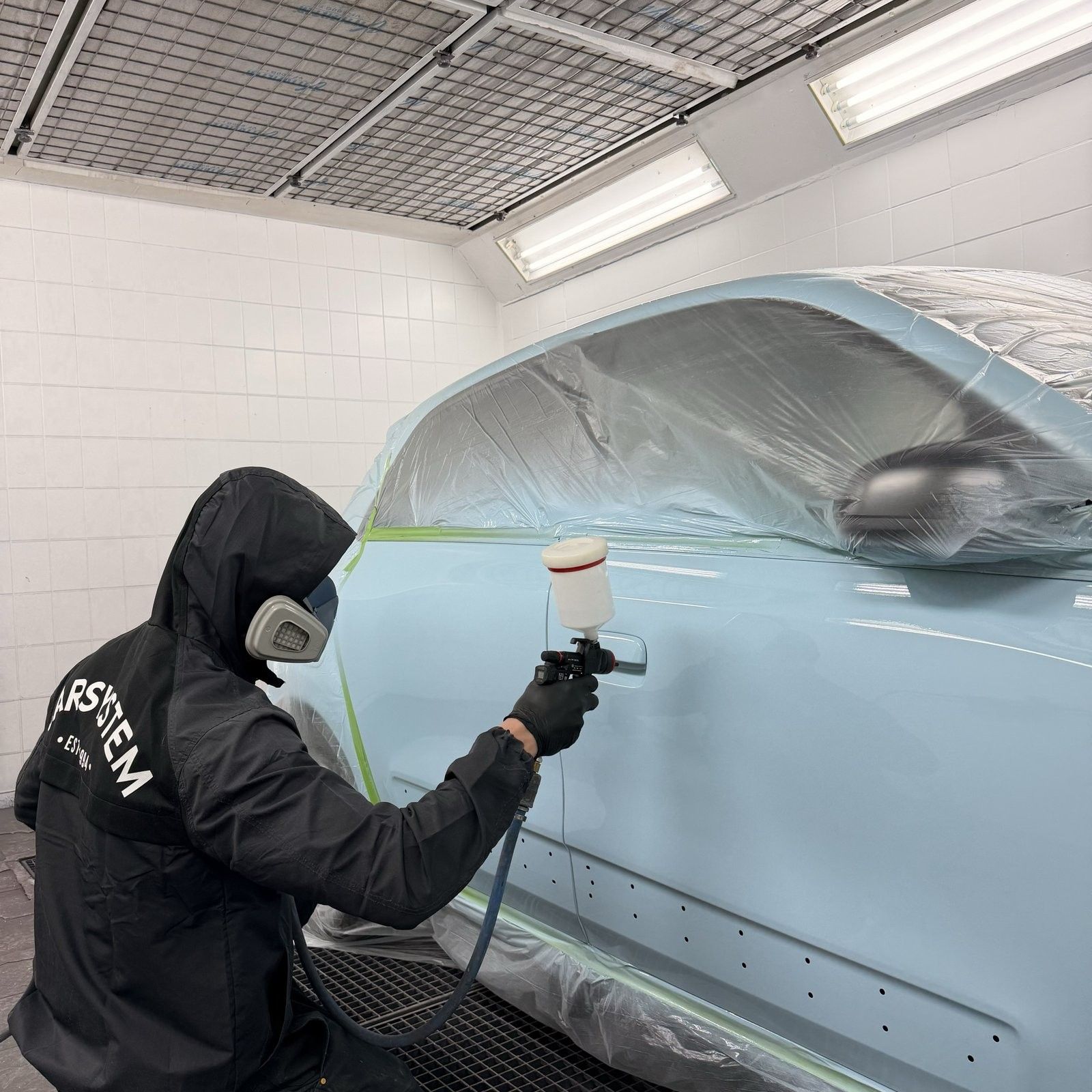 Lackierer trägt in der Lackierkabine Lack auf eine abgeklebte Fahrzeugseite auf